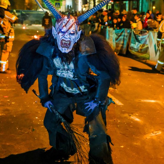 Eine Person in Teufelskostüm steht nachts auf einer Straße. Eine Menge beobachtet hinter Absperrungen. Das Kostüm besteht aus Hörnern, einer Maske und einem schwarzen Umhang.