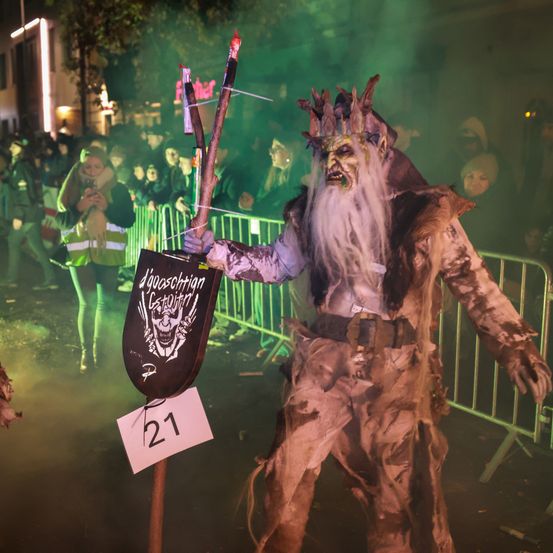 Eine Person in einer Zombie-Verkleidung steht auf einer Bühne, hält eine Lanze in der Hand, mit einer Menge, die hinter einer grünen Barriere zuschaut. Der Zombie hat ein Schild mit der Nummer 21.
