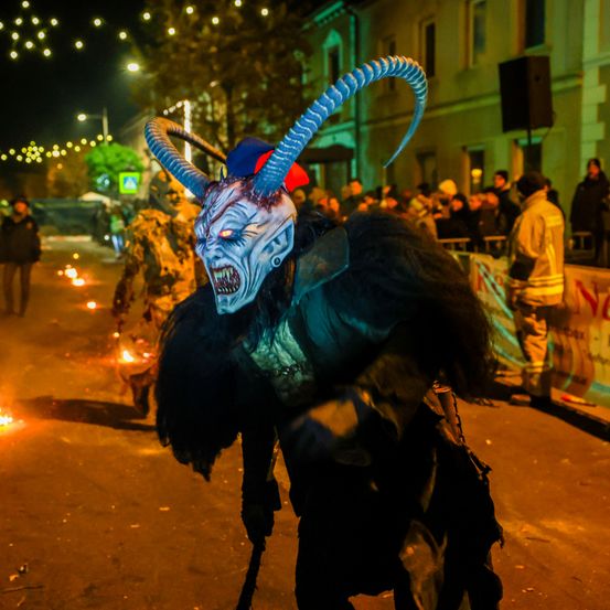 Eine Person in einem Dämonenkostüm mit Hörnern und Maske tritt bei Nacht auf einer Straße auf, während Feuerstellen und eine Menge zusehen.