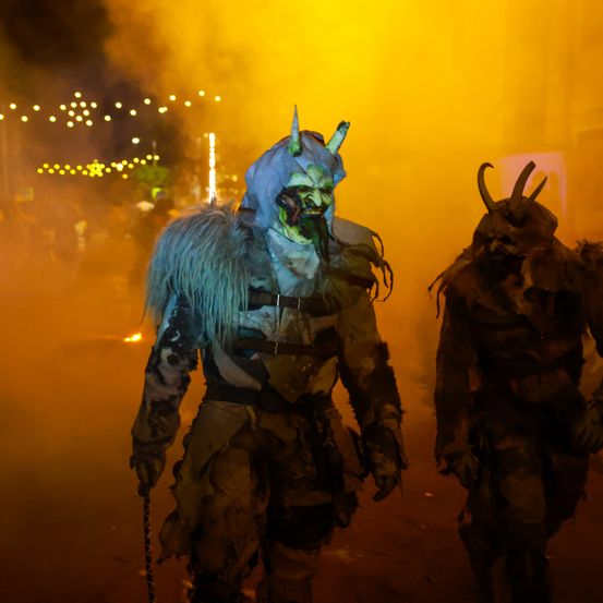 Zwei Personen in Hörnermasken gehen in einem orange-gelben Nebel mit Lichtern im Hintergrund.