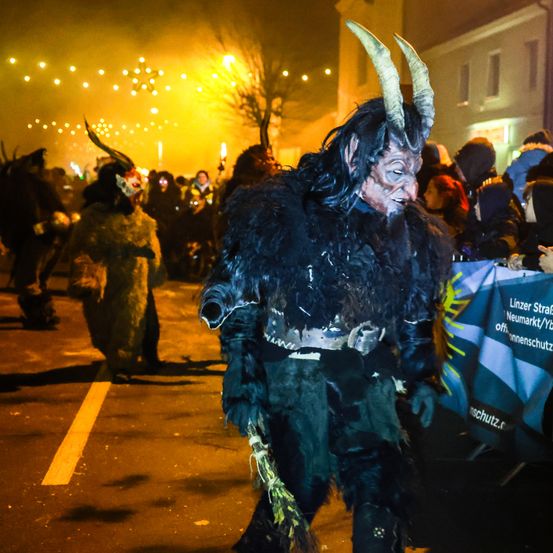 Eine Gruppe von Menschen in Hörnerkostümen zieht bei Nacht durch die Straße. Im Hintergrund sind brennende Kerzen und ein gelbes Leuchten zu sehen.