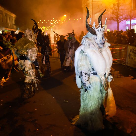 Eine Gruppe von Menschen in Hörnerkostümen geht nachts auf der Straße. Sie sind von einer Menge umgeben und in der Ferne gibt es Lichter und ein Feuer.