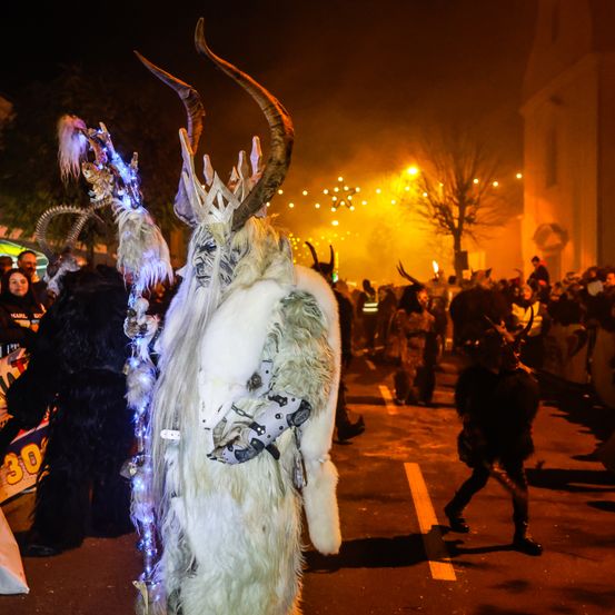 Eine Person in einem weißen Kostüm mit Hörnern und einer Maske geht bei Nacht bei einer Parade. Es sind Leute um ihn herum.