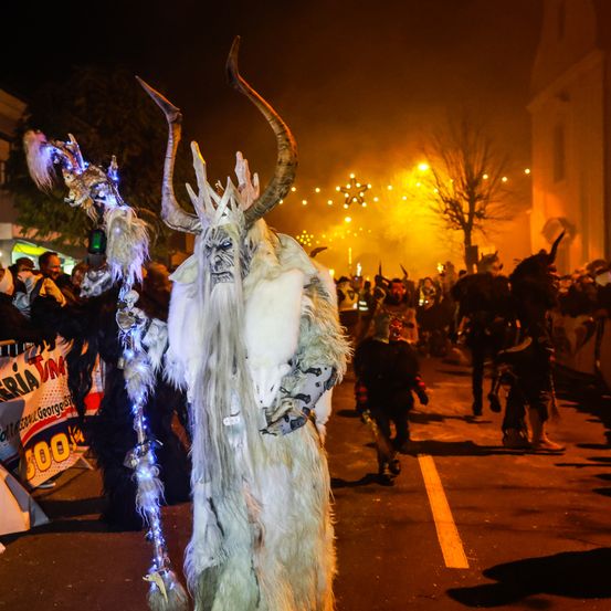 Eine Person, die als Schneemonster mit großen Hörnern verkleidet ist, geht in einer Straße mit anderen Menschen in Kostümen. Im Hintergrund sind Lichter und Bäume zu sehen.