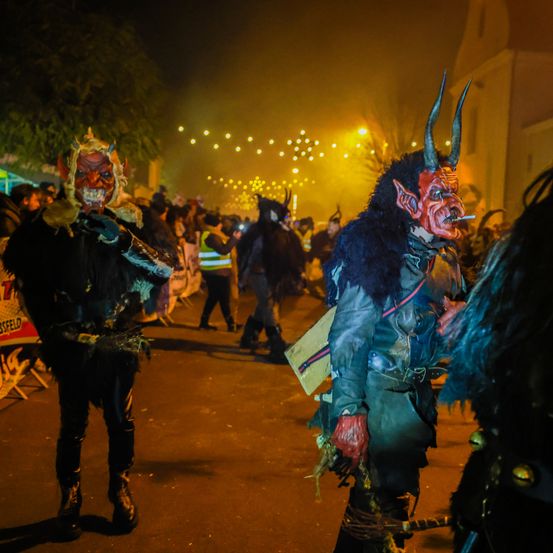Nachtfest mit Leuten in Teufelskostümen und Masken, die Zigaretten halten und auf einer Straße mit Straßenlaternen laufen.
