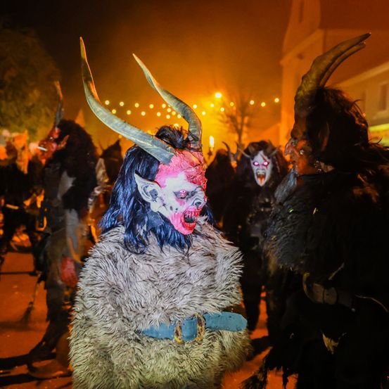 Eine Gruppe von Menschen in gehörnten Kostümen und Gesichtsmasken geht bei einem nächtlichen Festival zusammen. Die Kostüme sind pelzig und die Masken sind in Rot und Schwarz bemalt.
