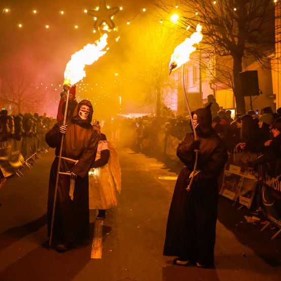 Zwei Personen in Kostümen mit Masken und brennenden Fackeln gehen bei Nacht in der Mitte einer Straße, umgeben von einer Menge.