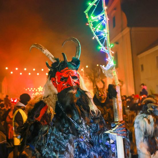 Ein Mann mit einer hörnigen Maske, der einen leuchtenden Stab bei einem nächtlichen Fest hält, umgeben von anderen maskierten Teilnehmern.