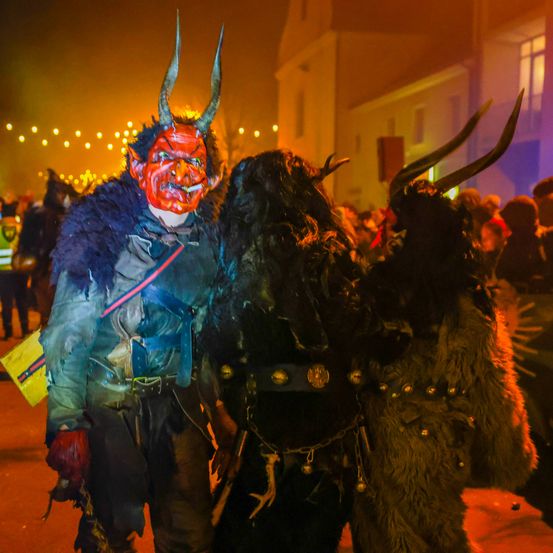 Zwei Personen in aufwendigen Kostümen, eine mit Hörnern und einer roten Maske, stehen zusammen auf einer schwach beleuchteten Straße. Der Hintergrund ist festlich mit einer Menge und Gebäuden.