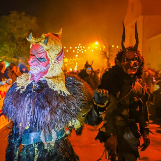 Zwei Personen in Hörnerkostümen gehen durch eine festliche, schwach beleuchtete Straße, umgeben von einer Menge, mit entfernten Lichtern und einem Gebäude im Hintergrund.