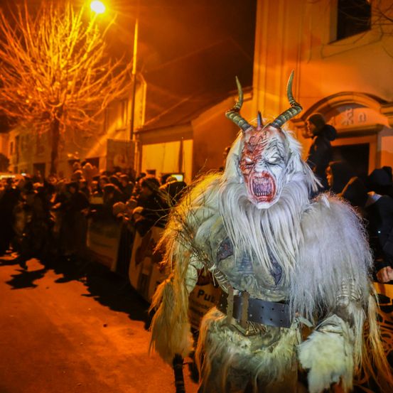 Eine Person in einem Dämonenkostüm mit Hörnern und einem weißen Bart geht bei Nacht auf der Straße, eine Menge schaut zu.