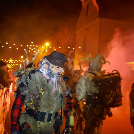 In der Nacht geht eine Person in einem gehörnten Kostüm vor einer Kirche mit rotem Rauch im Hintergrund.
