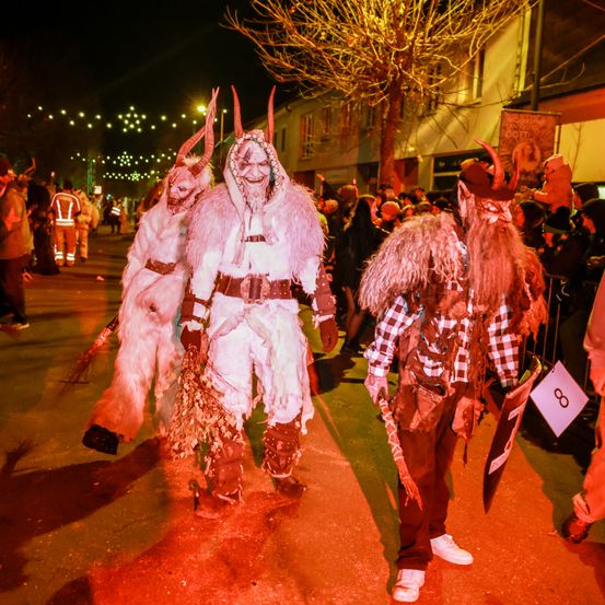 Drei kostümierte Personen mit Hörnern und Masken gehen bei Nacht auf einer Straße, die von roten Lichtern beleuchtet wird. Eine Menge beobachtet, wie sie die Straße hinuntergehen.