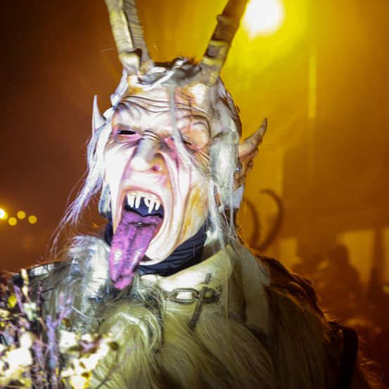 Eine Person mit einer Hörnermaske und einer violetten Zunge steht in einem schwach beleuchteten Bereich. Die Maske hat scharfe Hörner und einen finsteren Ausdruck. Der Hintergrund ist unscharf mit gelben und orangefarbenen Farbtönen.