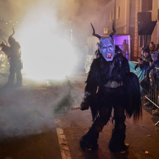 Eine Person, die als Dämon mit Hörnern verkleidet ist, bläst Rauch aus einer Pfeife vor einer Menge, wahrscheinlich während einer Halloween- oder Festivalveranstaltung. Dahinter ist eine weitere, ähnlich gekleidete Person zu sehen.