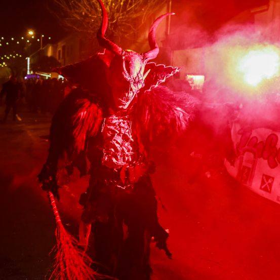 Eine Person in einem roten Teufelskostüm mit Hörnern und einem Schwanz läuft bei Nacht auf einer Straße mit einer Menschenmenge im Hintergrund.