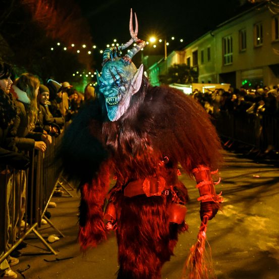 Eine Person in einem roten Kostüm mit Hörnern und einer Maske läuft nachts auf der Straße. Viele Menschen beobachten ihn hinter dem Zaun.