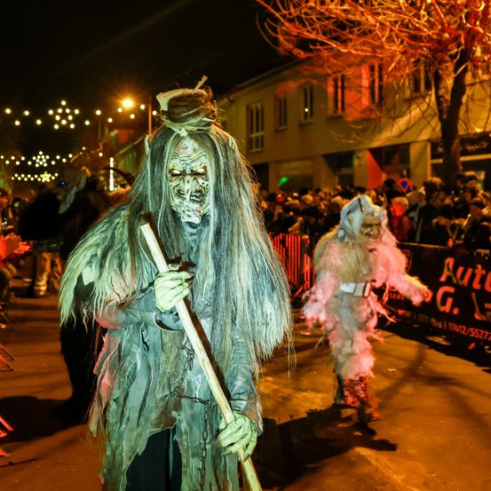 Zwei Personen in gruseligen Kostümen, möglicherweise für Halloween, gehen nachts mit Stöcken in der Straße. Eine Menge Leute beobachtet von hinten. In der Ferne stehen Gebäude und Bäume.