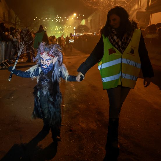 Eine Person in einem Kostüm geht bei Nacht die Straße entlang und hält die Hand einer Frau, die eine Warnweste trägt. Zuschauer beobachten von der Seite.