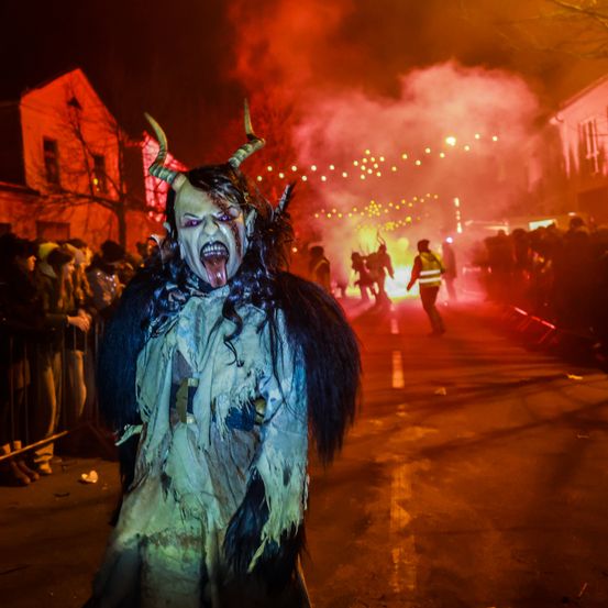 Eine Person in einem Teufelskostüm steht auf der Straße, umgeben von Rauch und einer Menschenmenge. Im Hintergrund befinden sich Menschen mit Hörnern und Feuerwerk.