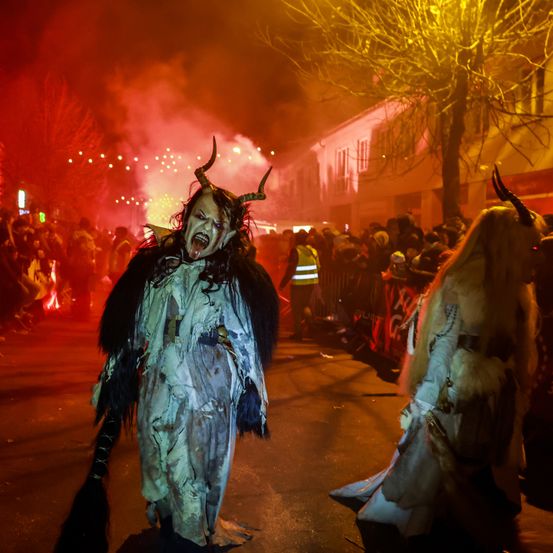 Zwei Personen in Hörnerkostümen gehen bei Nacht auf einer Straße, umgeben von einer Menge und roten Lichtern.