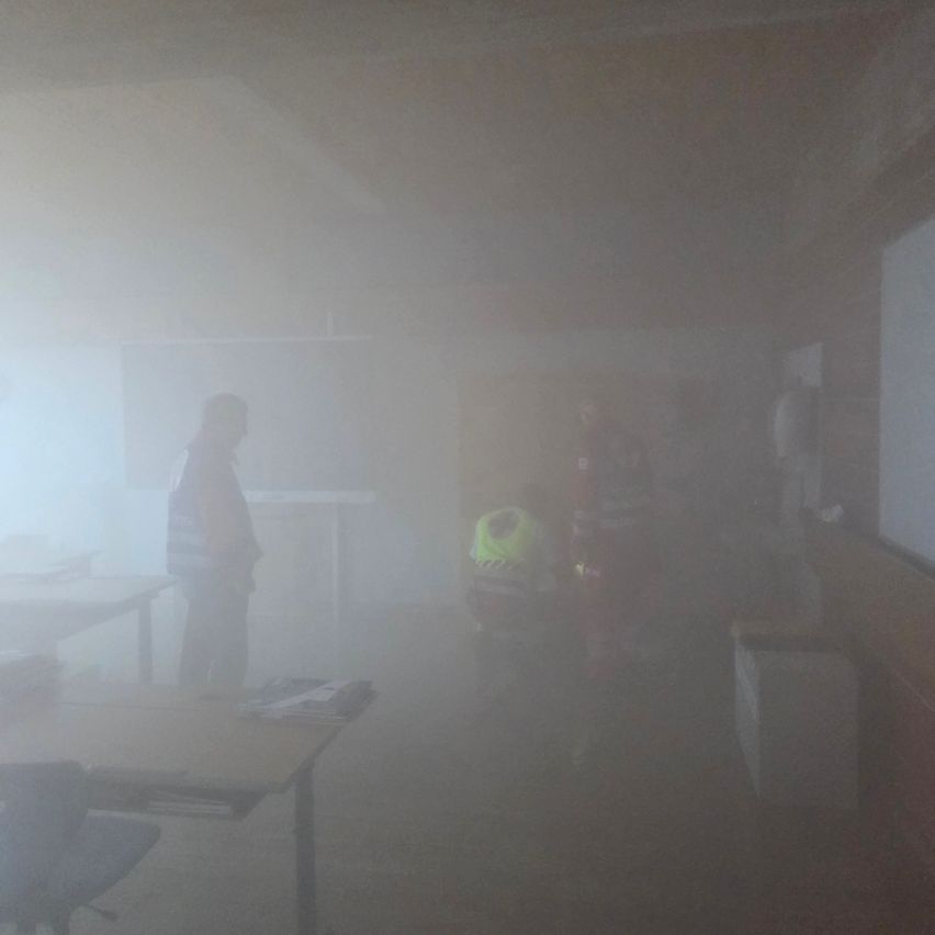 A foggy room with several people in safety gear, one kneeling on the floor. Tables and chairs are visible, but details are obscured by smoke.