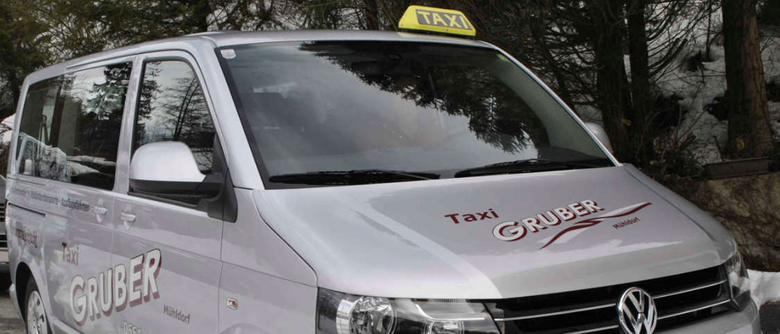 Ein silberner Volkswagen-Taxi-Van mit einem TAXI-Schild oben, dem Namen Gruber und der Nummer 0654-1144737 an der Seite. Der vordere Stoßfänger hat das Volkswagen-Logo.