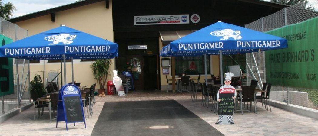 Der Außenbereich eines Restaurants namens Puntigamer mit blauen Sonnenschirmen, einer Küchenchef-Statue und einer Tafel.