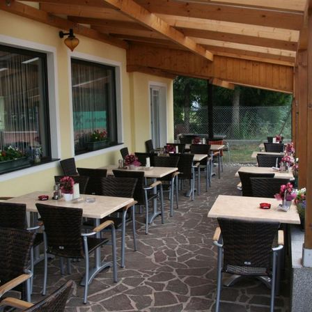 Ein Essbereich im Freien mit Holzbalken und Steinboden, blumengeschmückten Tischen und Stühlen, einer Lampe an der Wand und einem grünen Zaun mit Bäumen.