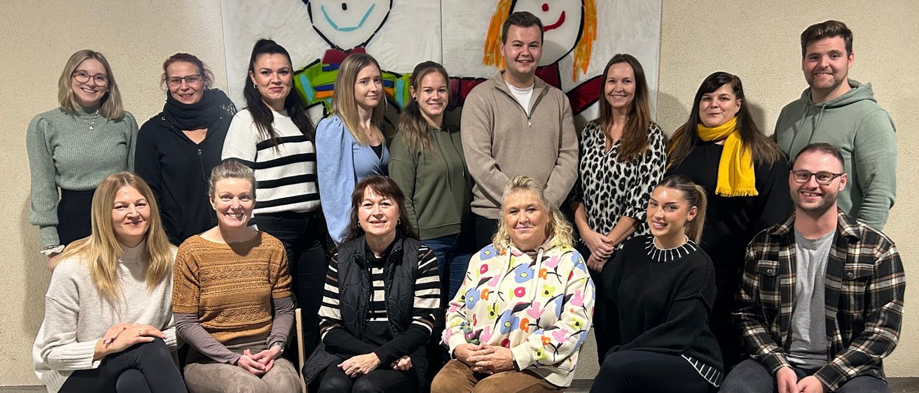 Eine Gruppe von Menschen posiert für ein Foto in einem Raum mit zwei großen Zeichnungen eines Jungen und eines Mädchen an der Wand. Sie sitzen und stehen auf Stühlen und lächeln.