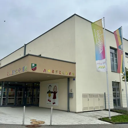 Der Eingang eines Kindergartens mit dem Namen 'Ansfelden'. Fahnen sind an Stangen neben dem Gebäude angebracht. Das Gebäude hat große Fenster und eine Wandmalerei von Kindern.