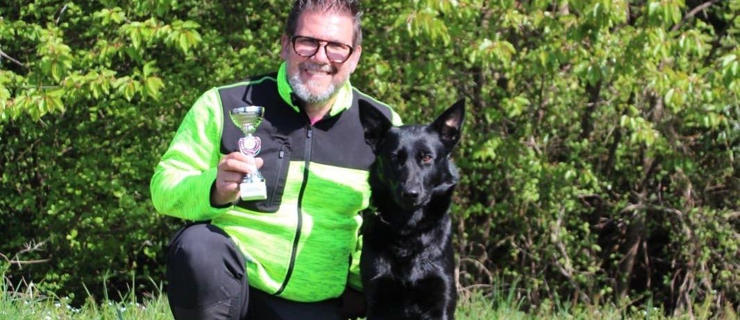 Ein Mann mit Brille hält eine Trophäe, während er mit seinem schwarzen Hund auf einem Feld kniet.