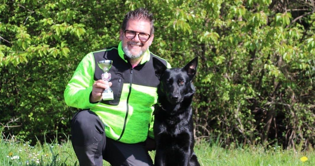Ein Mann mit Brille hält eine Trophäe, während er mit seinem schwarzen Hund auf einem Feld kniet.