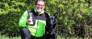 Ein Mann mit Brille hält eine Trophäe, während er mit seinem schwarzen Hund auf einem Feld kniet.