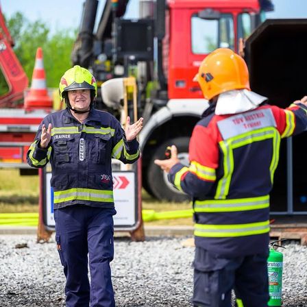 Zwei Feuerwehrleute in Schutzkleidung, einer erklärt und gestikuliert mit den Händen, der andere hört zu, stehen vor einem Feuerwehrauto und Sicherheitskegeln.