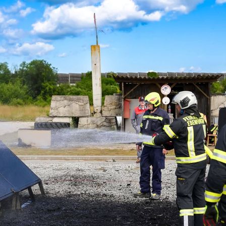 Feuerwehrleute üben mit einem Wasserwerfer, der Wasser auf verbranntes Material sprüht. Sie tragen Helme und Schutzanzüge. Hinter ihnen ist ein Gebäude mit einem Schild sichtbar.