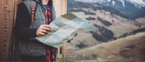 Eine Person hält eine Karte mit einer Berglandschaft im Hintergrund. Sie trägt eine Weste und ein kariertes Hemd.