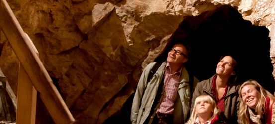 Eine vierköpfige Familie steht lächelnd in einer Höhle. Ein erwachsener Mann und eine Frau tragen Jacken. Ein junges Mädchen und ein Junge tragen rote und weiße Hemden. Treppen sind links.