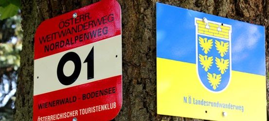Zwei Schilder an einem Baum, eines rot mit weißen und schwarzen Texten und das andere blau und gelb mit einem Wappen. Das rote Schild zeigt 'Osterreich. Weitwanderweg Nordalpenweg 01.' Das blaue Schild zeigt 'NO. Landesrundwanderweg.'