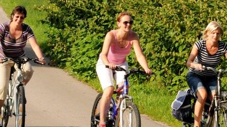 Drei Frauen fahren auf einem asphaltierten Weg mit Fahrrädern, mit Grünpflanzen und Büschen auf beiden Seiten. Sie tragen Freizeitkleidung und Sonnenbrillen.