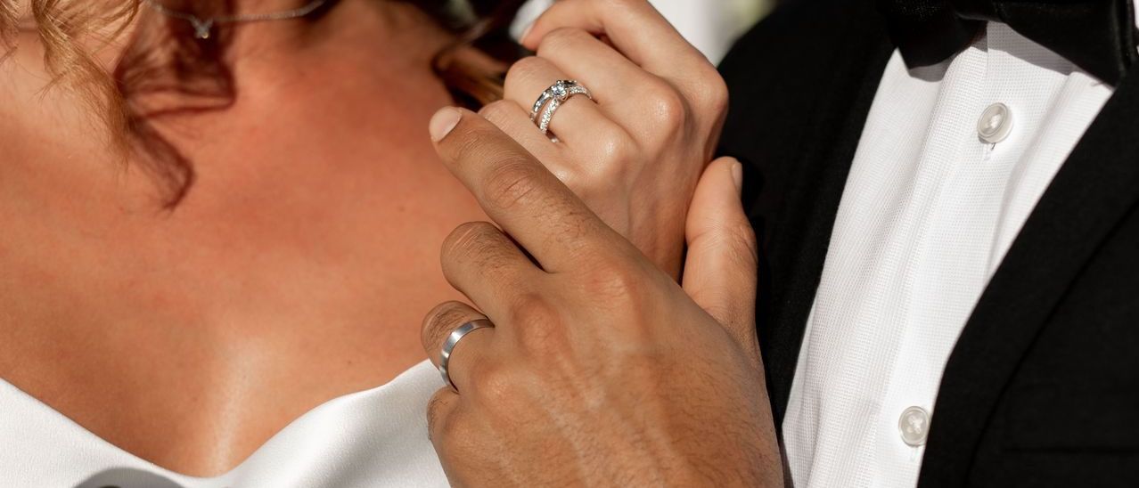 Eine Braut und ein Bräutigam halten Händchen, die Hand des Bräutigams liegt am Ringfinger der Braut, beide tragen Ringe.