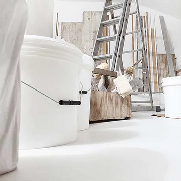 Ein Raum mit einem weißen Eimer, einer Leiter und einer Holzkiste mit Malutensilien auf dem Boden.