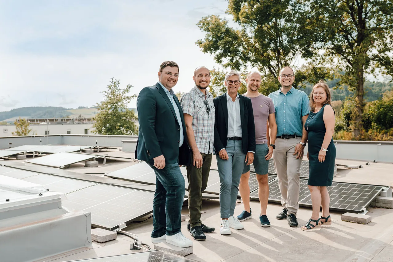 Sechs Personen, in unterschiedlicher Kleidung, stehen auf einem Dach mit Solarpanelen im Hintergrund, unter einem klaren Himmel.