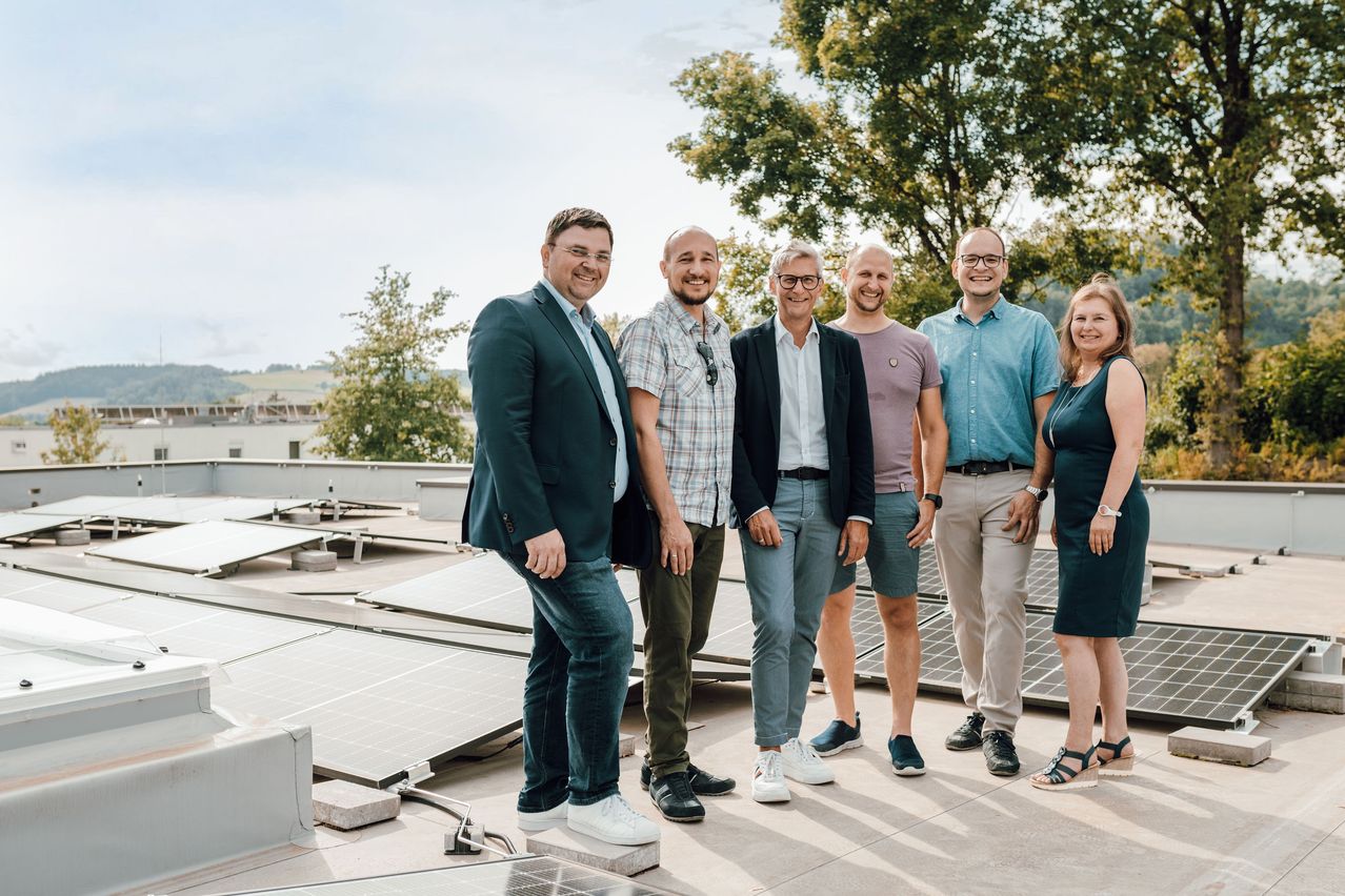 Sechs Personen, in unterschiedlicher Kleidung, stehen auf einem Dach mit Solarpanelen im Hintergrund, unter einem klaren Himmel.