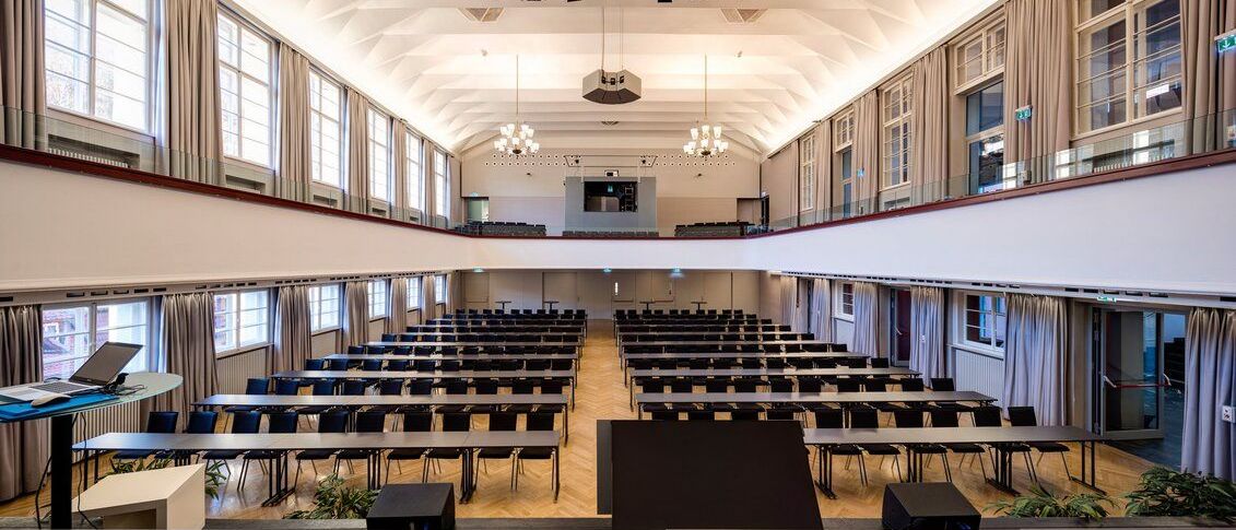 Ein leerer Konferenzraum mit Reihen von Tischen und Stühlen, einem Rednerpult, einer Projektionsleinwand und Kronleuchtern.