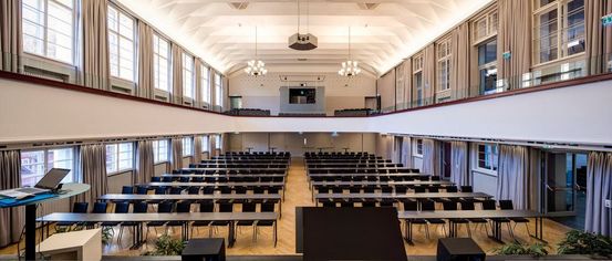 Ein leerer Konferenzraum mit Reihen von Tischen und Stühlen, einem Rednerpult, einer Projektionsleinwand und Kronleuchtern.