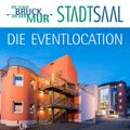 Ein modernes Gebäude mit einer Wendeltreppe, Glasfenstern und einem Schild mit der Aufschrift 'Die Eventlocation'. Es wird vom Abendsonnenschein beleuchtet, mit einem klaren blauen Himmel im Hintergrund.