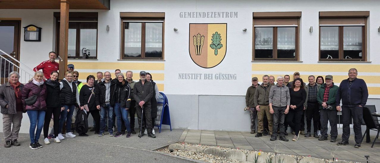 Several people stand in front of the Neustift bei Gussing community center, holding golf clubs. A yellow and brown emblem adorns the wall above them.