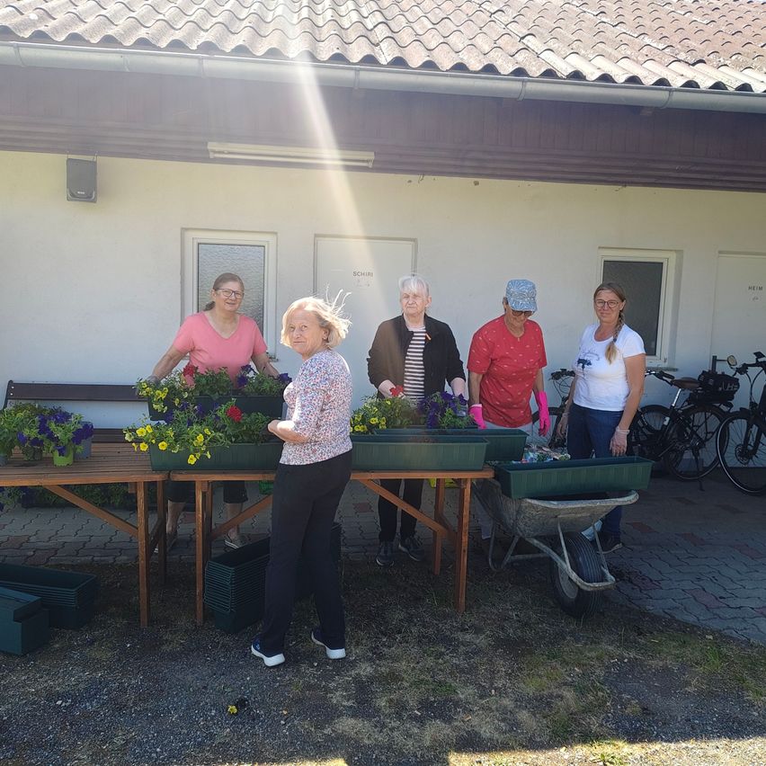 Five women are standing outside in front of a building. They are arranging flower pots on a table. One woman is pushing a wheelbarrow. The building has a white wall and a brown roof.