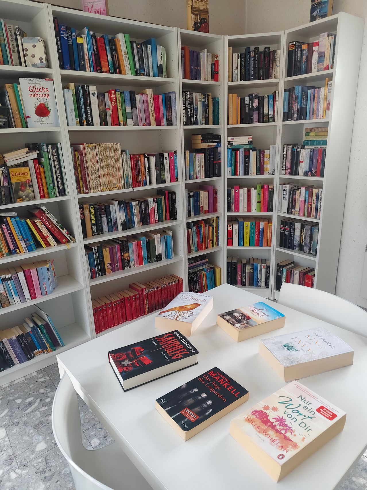 A well-stocked library with books on white shelves and a table with several books on it.